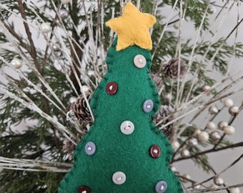 Può includere: Un ornamento per l'albero di Natale fatto a mano. L'albero di feltro verde è adornato con bottoni colorati e sormontato da una stella gialla. L'ornamento è sospeso da uno spago ed è posto su uno sfondo di rami sempreverdi ed elementi decorativi.