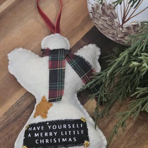 May include: A white felt angel ornament with a red velvet hanging loop, a plaid fabric scarf, a small gold star patch, and a black rectangular label featuring the white text "HAVE YOURSELF A MERRY LITTLE CHRISTMAS" with gold corner accents.