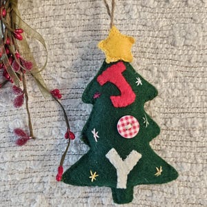 May include: A handmade Christmas tree ornament. The green felt tree is adorned with a red letter "J", a white letter "Y", a red and white checkered button, and small yellow star accents. A yellow star tops the tree.