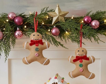 May include: Two tan felt gingerbread man ornaments hanging from a green garland. Each ornament features a red velvet bow, two colorful buttons, stitched facial details, and white rickrack trim on the arms and legs, suspended by red ribbons.