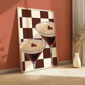 May include: A framed print of two coffee cocktails with a brown and white checkered background. The cocktails are in martini glasses and have a brown coffee base with a white foamy top. Coffee beans are visible on top of the foam.