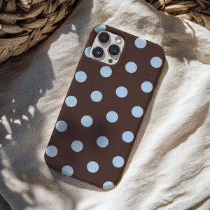 May include: A brown phone case with a pattern of light blue polka dots. The case is made of a smooth material and has a camera cutout. The phone case is lying on a beige fabric.