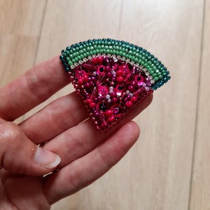 May include: A beaded watermelon slice brooch. The brooch features a green beaded rind, a white beaded layer, and a vibrant pink and red beaded interior with sequins. The brooch is a unique accessory.