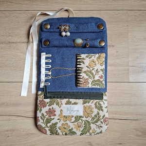 Può includere: Un astuccio portagioie in denim con una sezione inferiore a fantasia floreale e una piccola tasca con cerniera. Il rotolo presenta bottoni automatici color ottone e un nastro bianco. All'interno sono esposti diversi gioielli.