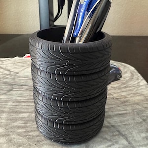 May include: A black tire pen holder with five stacked tires. The holder is filled with pens and pencils.