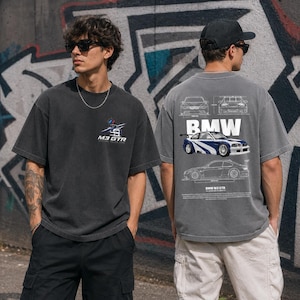 May include: A charcoal gray short-sleeve t-shirt featuring a small M3 GTR logo on the front chest. The back displays a large graphic of a blue and white BMW M3 GTR car with technical line drawings, the text BMW, and descriptive vehicle specifications.