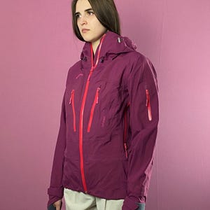 May include: A plum-coloured hooded jacket with red zips and accents. The jacket features multiple pockets and the word "lofoten" printed on the chest. The jacket is worn with light-coloured trousers.