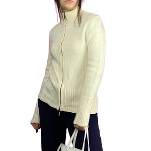 Puede incluir: Un cárdigan blanco de punto con cremallera, cuello alto y mangas largas.