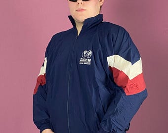 May include: Navy blue and red vintage track jacket with white accents. The jacket features a full zipper, a high collar, and a logo on the chest. The sleeves have red and white color blocks. The person is wearing black sunglasses and a silver watch.