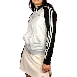 Puede incluir: Una sudadera con capucha azul claro con cremallera, mangas negras y tres rayas blancas en cada manga. La sudadera tiene un logotipo de Adidas negro en el pecho izquierdo.