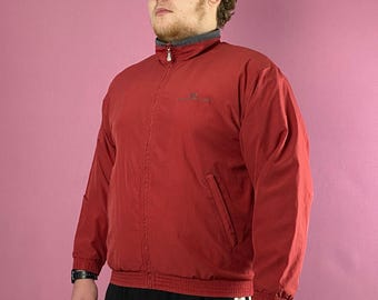Op de afbeelding: Rode jas met grijze kraag en klein logo op de borst. De jas heeft een ritssluiting en twee zakken. De persoon draagt een zwarte zonnebril en een zwart-witte trainingsbroek.