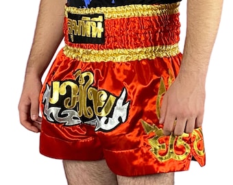 May include: Red and gold Muay Thai shorts with Thai lettering and gold trim. The shorts have a shiny, satin-like appearance and feature intricate gold and silver designs. The waistband is gold and has black text.