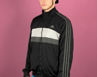May include: Black track jacket with white and gray horizontal stripes across the chest. The jacket has gray stripes down the sleeves and a small Adidas logo on the chest. The person in the image is wearing a purple baseball cap.