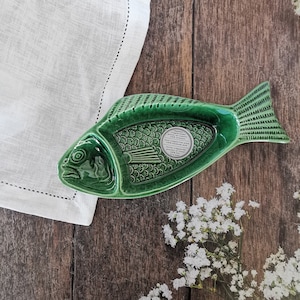 May include: A green ceramic fish-shaped serving dish with detailed scales and fins. The dish is on a wooden surface next to a white linen napkin and small white flowers. The dish has a label with text.