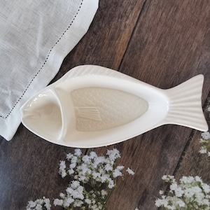 May include: White ceramic fish-shaped serving dish with a separate compartment for dipping sauce. The dish has a textured surface that resembles fish scales.