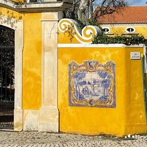 May include: A yellow gate with white trim and a black metal gate. The gate is decorated with a blue and white tile mural depicting a scene with trees and a building. The gate is surrounded by a yellow wall with white trim.