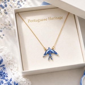 May include: A gold chain necklace with a swallow-shaped pendant featuring a blue and white floral pattern. The necklace is presented in a white gift box with the text Portuguese Heritage printed in gold on the inner lining of the box.