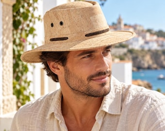 May include: A tan cork fedora hat with a flat crown and a wide brim. The hat features two black metal ventilation eyelets on each side and a dark brown horizontal band detail encircling the base of the crown, providing a textured appearance.