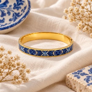 May include: A gold-toned hinged bangle bracelet featuring a central band with a blue and white floral tile pattern. The bracelet has a polished metallic interior and a secure clasp mechanism, showcasing a repeating intricate decorative design.