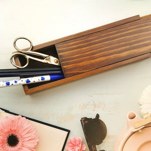 Op de afbeelding: Een houten potlooddoos met een scharnierend deksel, open om een ​​paar zilveren scharen, een zwart potlood en een blauw-witte pen te laten zien. De doos staat op een wit oppervlak.