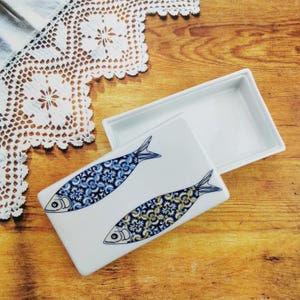 Op de afbeelding: Een rechthoekige witte keramische doos met een deksel, versierd met twee blauwe en gele vissen met ingewikkelde patronen. De doos staat op een houten oppervlak.