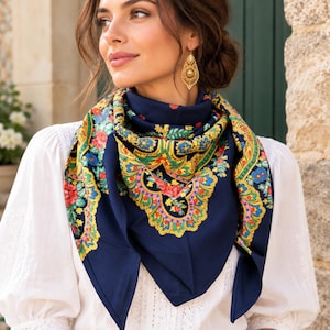 Puede incluir: Un pañuelo cuadrado de color azul marino con un vibrante estampado floral en tonos rojos, amarillos, verdes y azules, sobre una blusa blanca de manga larga. El pañuelo presenta bordes intrincados de estilo cachemir y motivos botánicos detallados.