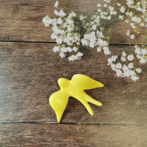 May include: A bright yellow ceramic bird figurine with a simple design, perched on a rustic wooden surface with white flowers in the background.