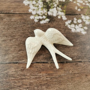May include: White ceramic swallow figurine with detailed wings. The figurine is in flight with its wings spread wide.