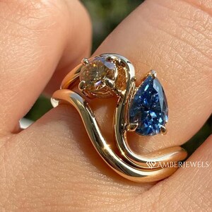 May include: A gold ring with a unique design, featuring two gemstones. One is a round, light brown stone, and the other is a pear-shaped, blue gemstone. The ring has a flowing, wave-like band. The text "AMBERJEWELS" is visible.