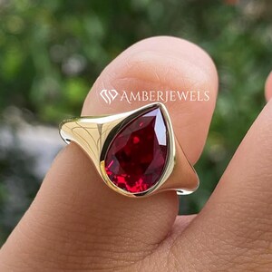 Puede incluir: Un anillo de oro con una piedra preciosa roja oscura en forma de pera. El anillo tiene una banda lisa y pulida y un engaste de bisel. La marca "AMBERJEWELS" es visible en el anillo. La piedra es de color rojo intenso.