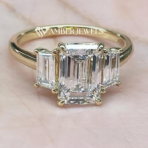 Puede incluir: Un anillo de oro con tres piedras preciosas de talla esmeralda. La piedra central está flanqueada por dos piedras más pequeñas, todo engastado en una banda de oro. El texto "AMBER JEWELS" es visible en el anillo. El anillo está sobre una superficie rosa.
