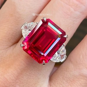 Op de afbeelding: Een opvallende ring met een grote, smaragdgeslepen, levendige rode edelsteen, geflankeerd door twee peervormige, heldere stenen. De ring is gezet in een zilverkleurig metaal. De merknaam "AMBERJEWELS" is zichtbaar.