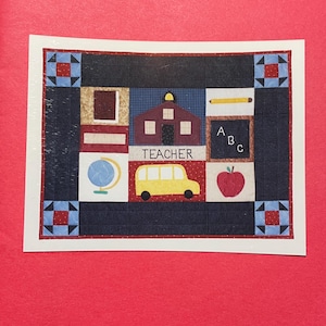 May include: A white card with a colorful patchwork quilt design featuring a schoolhouse, apple, globe, bus, and chalkboard with the letters ABC. The text "TEACHER" is printed on the quilt. The card has a red border and the text "Apple for the Teacher" and "SEAGULL QUILTS" are printed on the card.