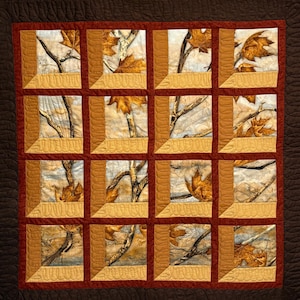 May include: A quilted blanket featuring a repeating pattern of square panels. Each panel showcases a nature scene with autumn leaves and tree branches. The panels are framed in a golden-brown color, with a dark brown border.