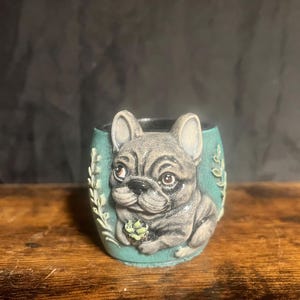 May include: A small, teal-colored ceramic pot with a detailed relief of a gray French Bulldog. The dog is centered on the pot, with floral accents on either side. The pot is sitting on a wooden surface.