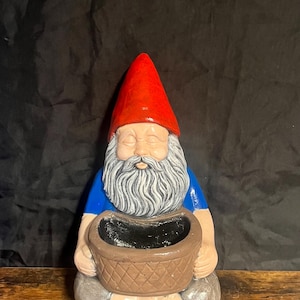 Puede incluir: Una figurita de gnomo de cerámica con un sombrero rojo, una camisa azul y una barba blanca. El gnomo sostiene una cesta marrón en su regazo.
