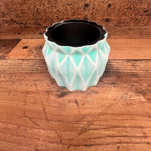 May include: A teal ceramic planter with a geometric diamond pattern. The planter has a black interior and a white glaze.