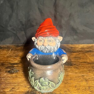 Pode incluir: Estatueta de gnomo de jardim em cerâmica com chapéu vermelho, camisa azul e barba branca, segurando um pote marrom com detalhes de folhas e rãs verdes. O gnomo tem um rosto detalhado e é um item decorativo de jardim.