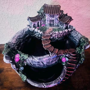 May include: A three-tiered ceramic planter with a small house on top. The planter is painted in shades of grey, brown, and green, with pink accents. The house has a brown roof and a grey exterior.