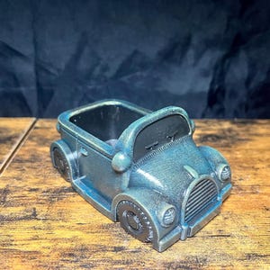 May include: A vintage-style car-shaped planter in shades of blue and gray. The planter has a detailed design, including headlights, a grill, and wheels. It is made of a textured material and sits on a wooden surface.