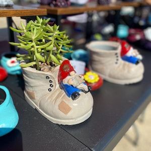 Puede incluir: Dos botas de cerámica blanca con una figura de gnomo sentada en cada una. Las botas están decoradas con un parche marrón y cordones negros. El gnomo en primer plano lleva un sombrero rojo, pantalones azules y barba blanca. Las botas se utilizan como macetas y contienen suculentas verdes.