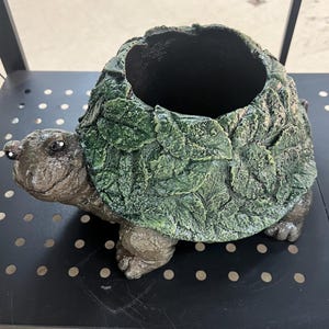 Puede incluir: Un macetero con forma de tortuga verde y marrón con un diseño de hojas. El macetero tiene una abertura ancha en la parte superior para plantar.