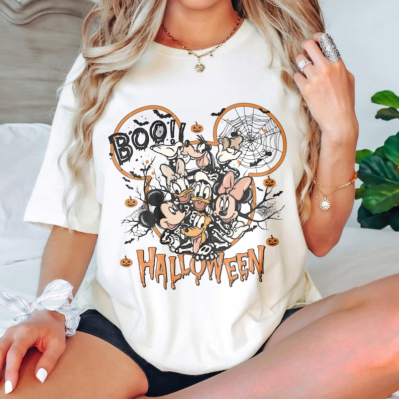 May include: White t-shirt with a Mickey Mouse Halloween design. The design features Mickey Mouse, Minnie Mouse, Donald Duck, Goofy, and Pluto dressed in Halloween costumes. The text "BOO!" and "HALLOWEEN" are also included in the design.