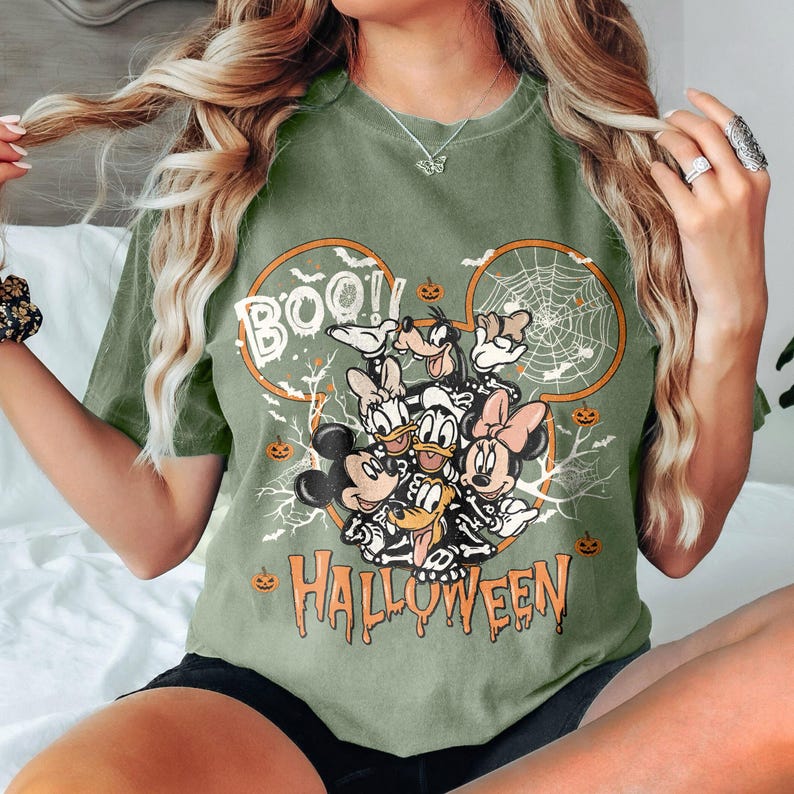 May include: Green t-shirt with a Mickey Mouse Halloween design. The design features Mickey Mouse, Minnie Mouse, Donald Duck, Goofy, and Pluto dressed in Halloween costumes. The text "BOO! HALLOWEEN" is printed on the shirt.