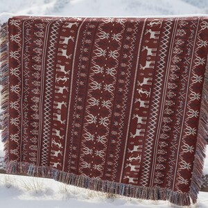 May include: A burgundy throw blanket with a white geometric pattern of reindeer and snowflakes hangs on a wooden fence covered in snow. The blanket has a fringed edge and is displayed outdoors with a snowy mountain backdrop.
