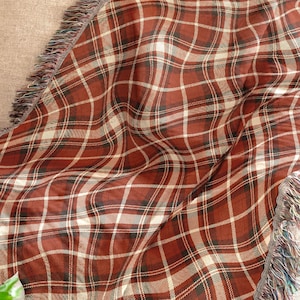 May include: A woven throw blanket with a plaid pattern in shades of red, white, and green. The blanket has a fringed edge and is draped over a beige sofa. The blanket is a cozy home decor item.
