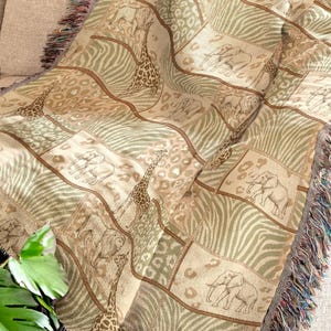 Puede incluir: Una manta tejida con temática de safari africano. La manta presenta un diseño de patchwork con imágenes de jirafas, elefantes y cebras en tonos beige, marrón y verde. Los bordes están acabados con un ribete de flecos.