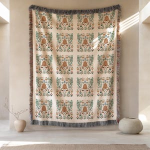 Puede incluir: Una manta de color crema con un patrón repetido de colmenas marrones, follaje verde y pequeños acentos florales rojos y azules. La manta tiene un borde con flecos en tonos grises y marrones, adecuada para una sala de estar o dormitorio.
