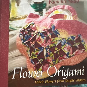 May include: A book titled "Flower Origami: Fabric Flowers from Simple Shapes" by Kumiko Sudo. The cover features a pink fabric bag with a colorful origami flower design.