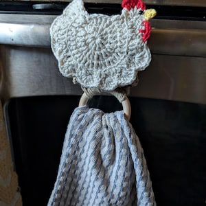 May include: A handmade crochet kitchen towel holder shaped like a chicken. The chicken is off-white with red and yellow accents. A blue and white striped towel hangs from a wooden ring attached to the chicken.